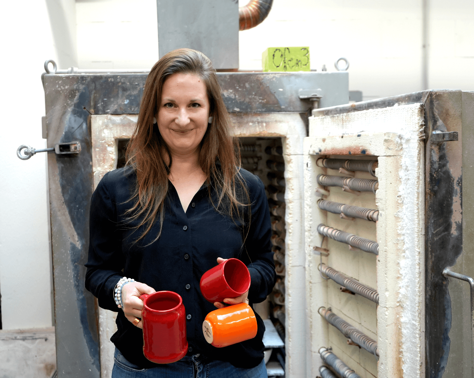 A woman with long brown hair, wearing a black blouse and jeans, stands in front of an open kiln, holding two glazed ceramic cups – one red and one orange – and a finished red jug. The open kiln reveals heating coils and shelves inside.