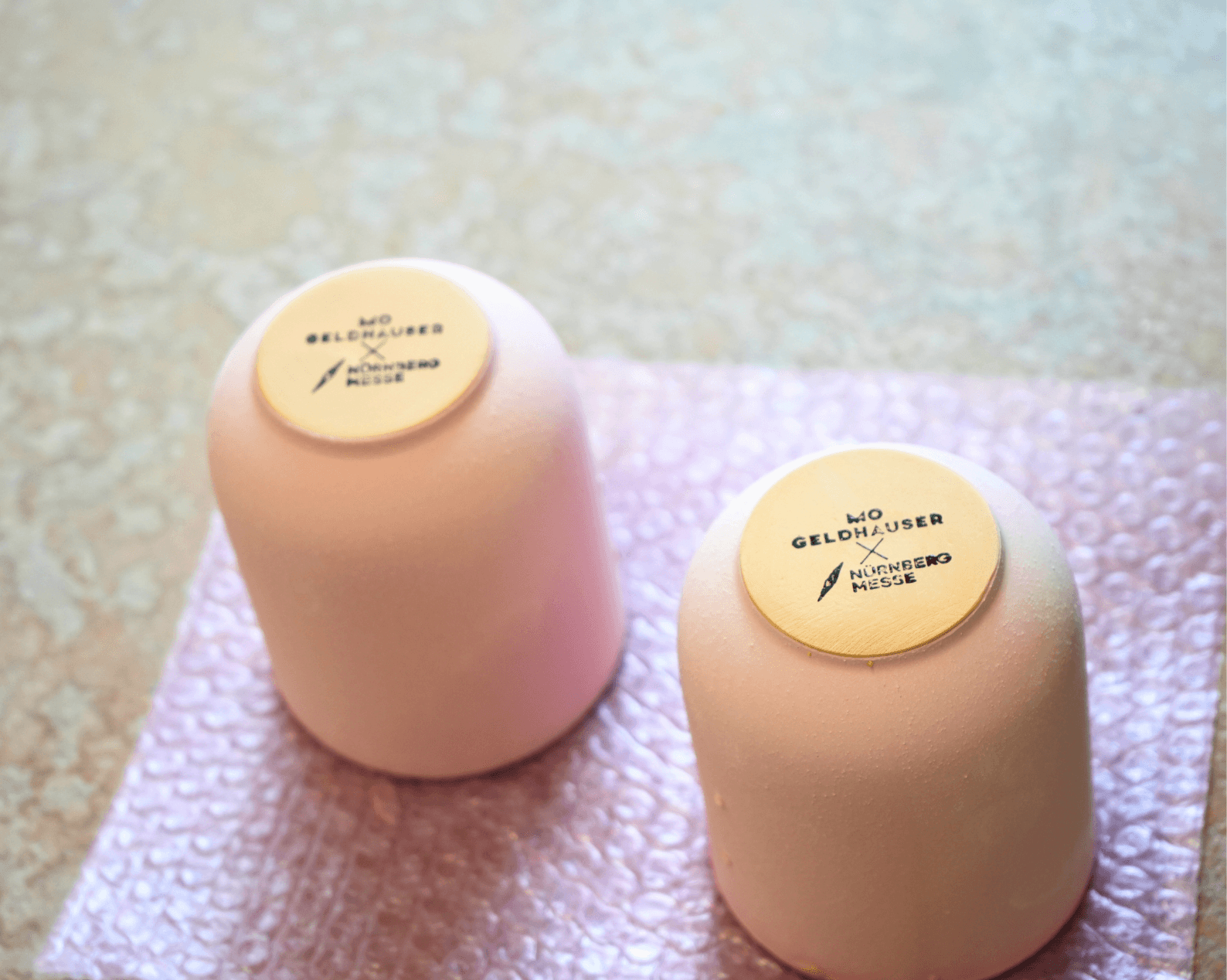 Two upside down pink ceramic cups rest on a padded surface. Circular labels on the bases read “Geldhauser × NürnbergMesse.”