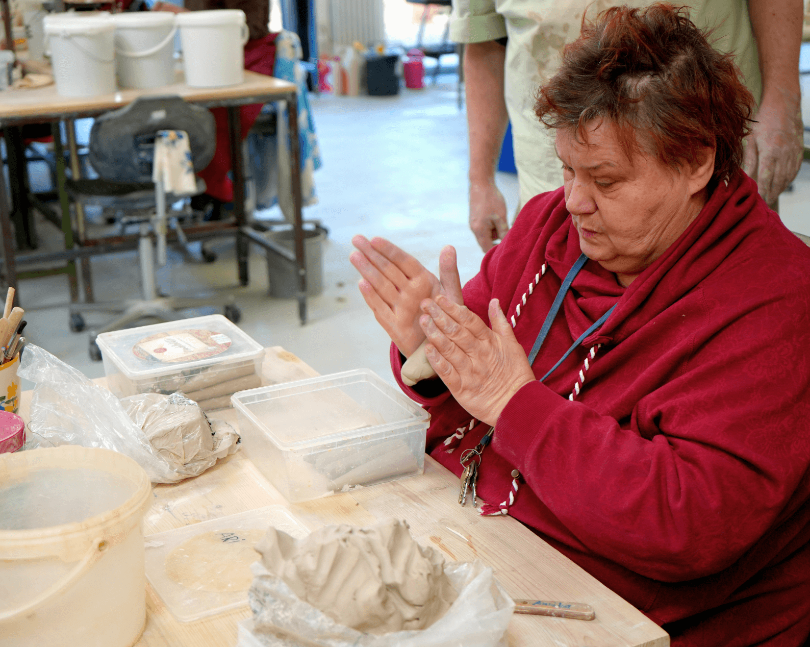 A woman with short black hair and orange highlights, wearing a red sweater, is sitting at a workbench, shaping clay with her hands. On the table are lumps of clay, toolboxes, plastic containers, and other materials used in pottery-making. In the background, workbenches and other workshop equipment are visible.