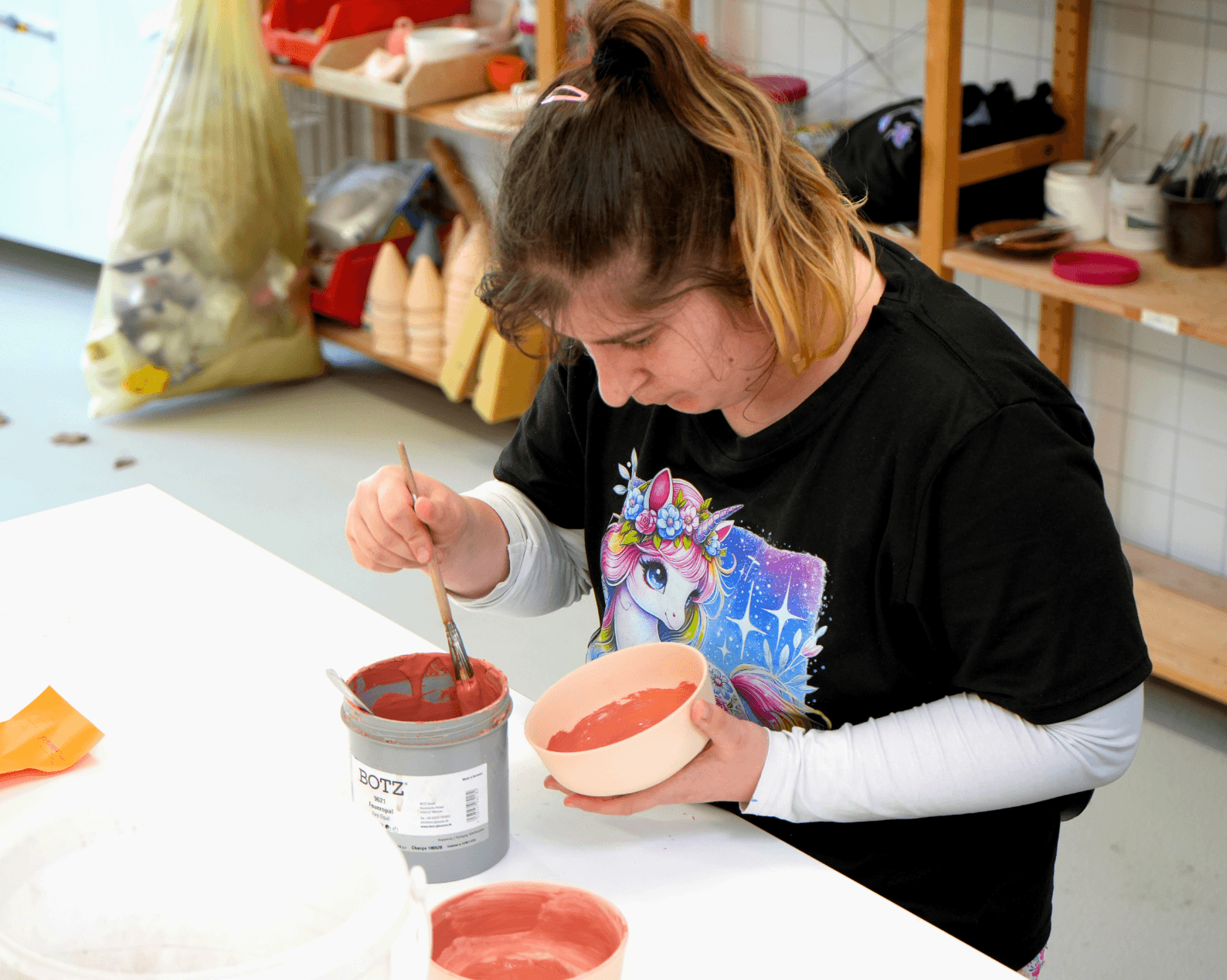 A woman with dark hair and a long blonde braid, wearing a white sweater and a black T-shirt printed with a unicorn, is sitting at a table and applying red glaze to a ceramic bowl with a brush. In front of her are glaze containers, tools, and other materials. In the background are shelves with supplies and packaging materials.