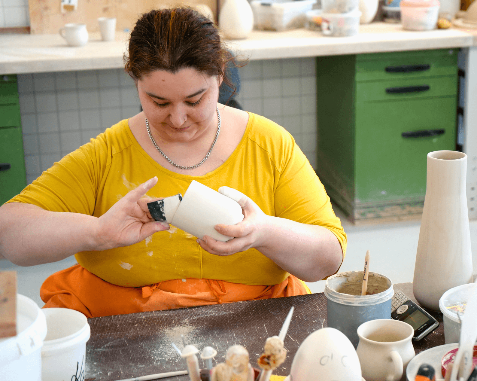 A woman with long brown hair, wearing a yellow T-shirt and an orange apron, is sitting at a workbench in a pottery studio, shaping a light-colored ceramic vessel with a tool. Various supplies are scattered across the table, including brushes, leftover clay, glaze containers, water jugs, and a cell phone. In the background is a work surface with additional unglazed ceramic pieces and tools.