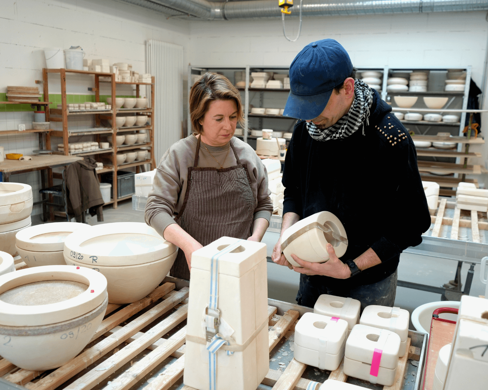 A woman and a man are standing in a ceramics workshop at a large workbench covered with numerous plaster molds, half-finished bowls, and other ceramic pieces. The woman with short blonde hair is wearing a brown sweater and a brown apron and is standing in front of several large round molds, while the man with short black hair, a cap, and a black-and-white scarf, dressed in a black sweater and jeans, is holding a plaster mold in his hands. In the background are shelves filled with additional ceramic molds and vessels, as well as workshop equipment.