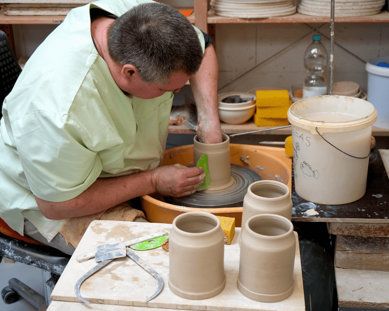 A man with short dark hair wearing a green T-shirt is working at a potter's wheel, shaping a cylindrical vessel out of clay. One of his arms guides a tool against the rotating form, while several other turned blanks, a bucket of water, and various tools lie on the workbench in front of him. In the background, shelves with additional ceramic pieces and a water bottle can be seen.
