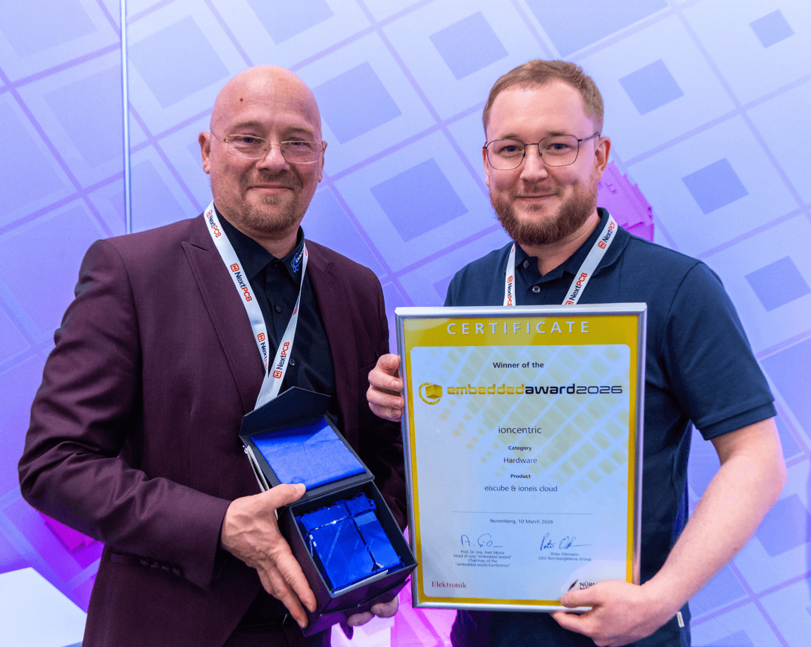 Der Mann links hält eine Glastrophäe, der Mann rechts hält ein gerahmtes Zertifikat.