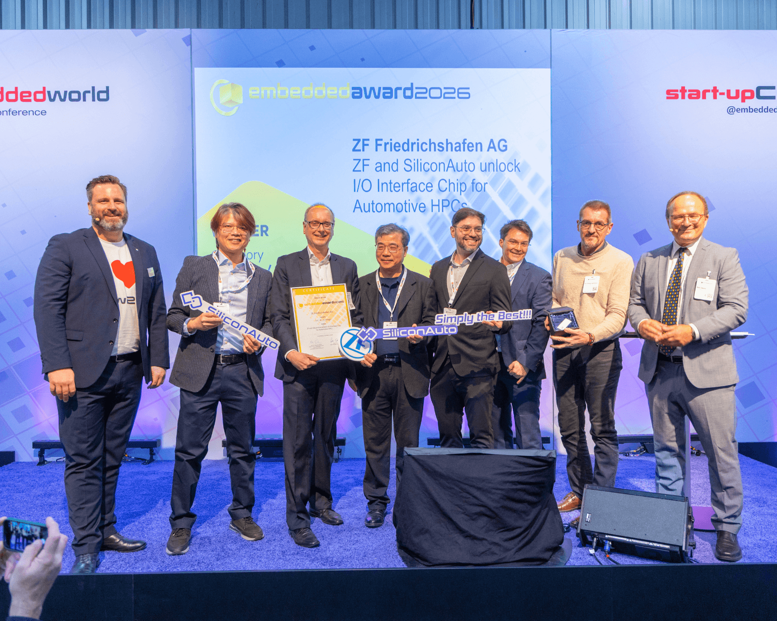 Acht Männer in Businesskleidung mit Zertifikat und Glastrophäe auf der Award Bühne.