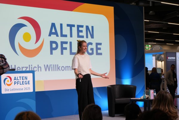 A woman stands on a stage on which “ALTENPFLEGE” is written in large letters. 