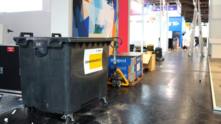 Müll-Recycling Ein großer schwarzer Müllcontainer mit der Aufschrift "Gewerbeabfall" steht in einem Messe- oder Ausstellungsbereich. Im Hintergrund sind bunte, geometrische Wanddekorationen und verschiedene Stände zu sehen. Eine Leiter und mehrere Kisten sowie andere Ausrüstungen sind ebenfalls im Bild sichtbar.