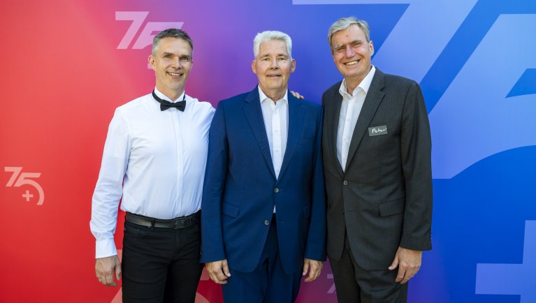 Three men in formal dress stand next to each other in front of a colored background that runs from red to blue. The man on the left is wearing a white shirt with a black bow tie and dark trousers, the man in the middle is wearing a dark blue suit with a white shirt, and the man on the right is wearing a dark suit with a name badge on the lapel. The numbers "75" and "75+" can be seen in the background.